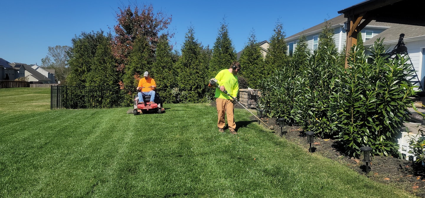 Two men trim a lush residential yard with a lawnmower and string trimmer.