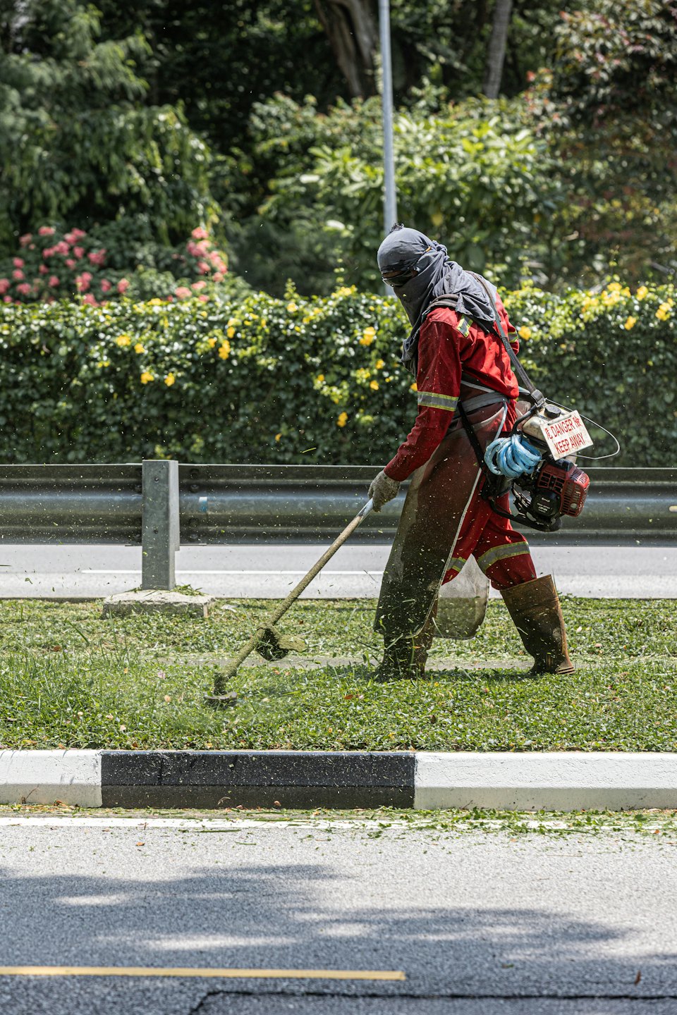 Worker in protective gear uses a trimmer along the roadside.